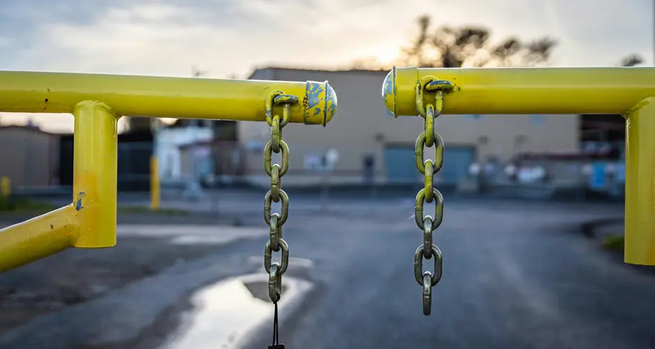 Gelber Absperrbügel mit zwei herabhängenden Ketten vor unscharfer Industriefläche.