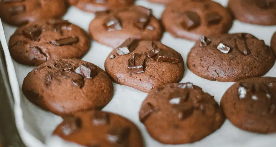 Mehrere schokoladenbraune Kekse mit Schokostückchen auf Backpapier, auf einem Blech gereiht.