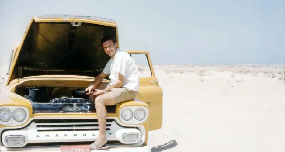 Mann sitzt am offenen Motor eines gelben Chevrolet in karger Wüstenlandschaft; Kühlergrill mit CHEVROLET-Schriftzug.