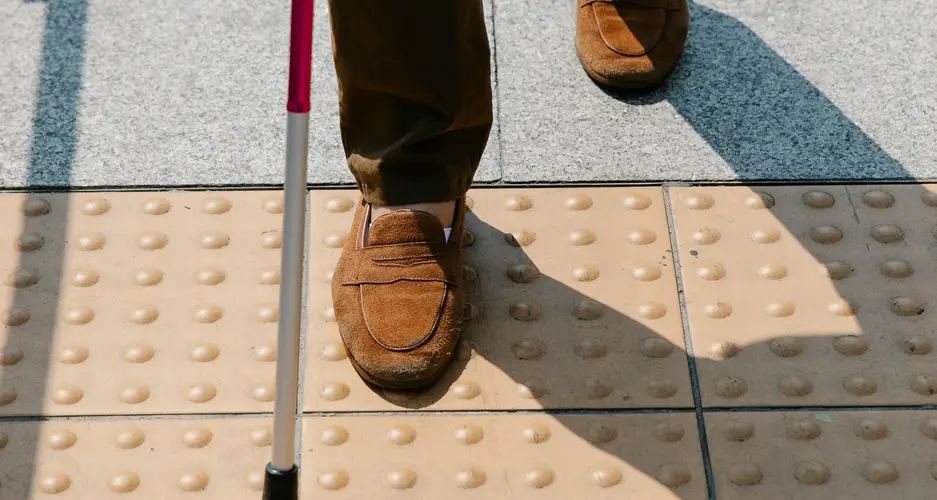 Person geht mit weiß-rotem Langstock über taktile Noppen als Leitsystem.