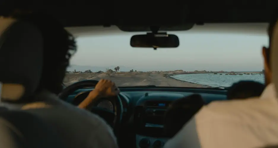 Blick aus dem Fond: Fahrer hat Hände am Lenkrad, Beifahrer rechts; Fahrzeug fährt auf schmaler Küstenstraße am Meer.
